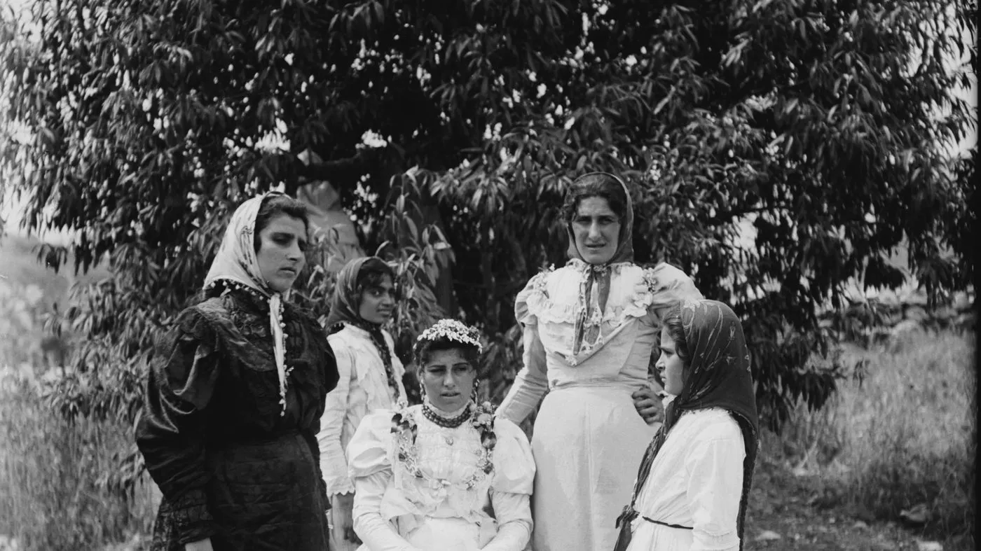 A wedding party of five women and girls photographed outdoors beneath a tree, the bride seated at the center in an embroidered white dress.