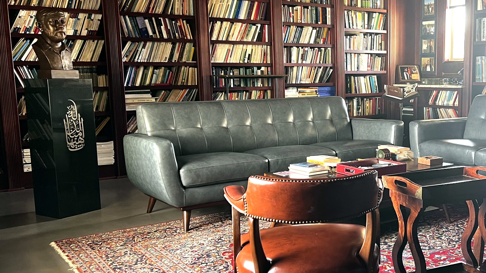 The reading room: walls of shelved books in dark wood cases, a leather sofa and chairs on a patterned carpet, and a bronze portrait bust on an inscribed plinth at the left.