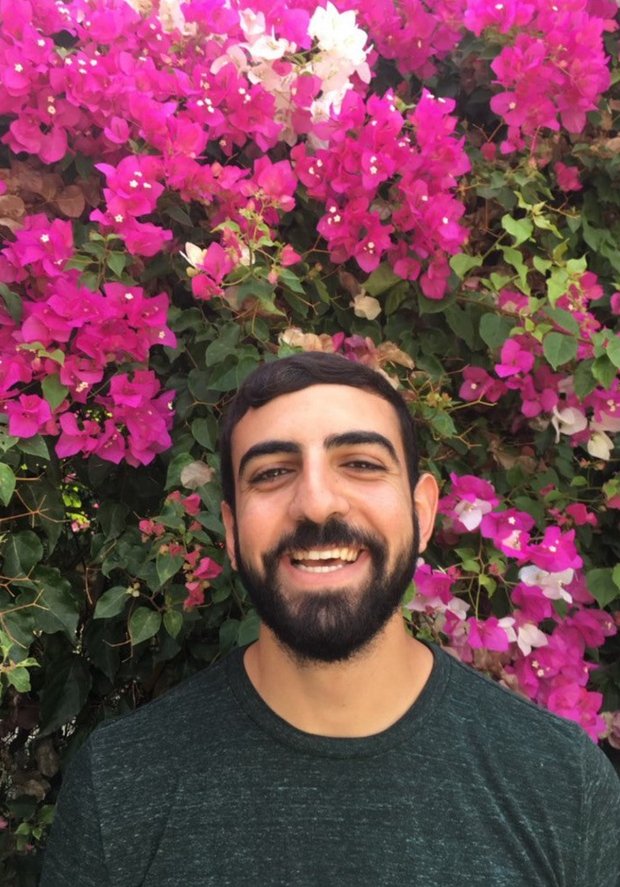 Senan Shaibani, a young bearded man, smiling in front of a flowering bougainvillea.